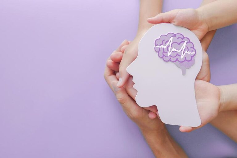 Hands holding paper cutout of head with purple brain and heartbeat symbol, representing brain health